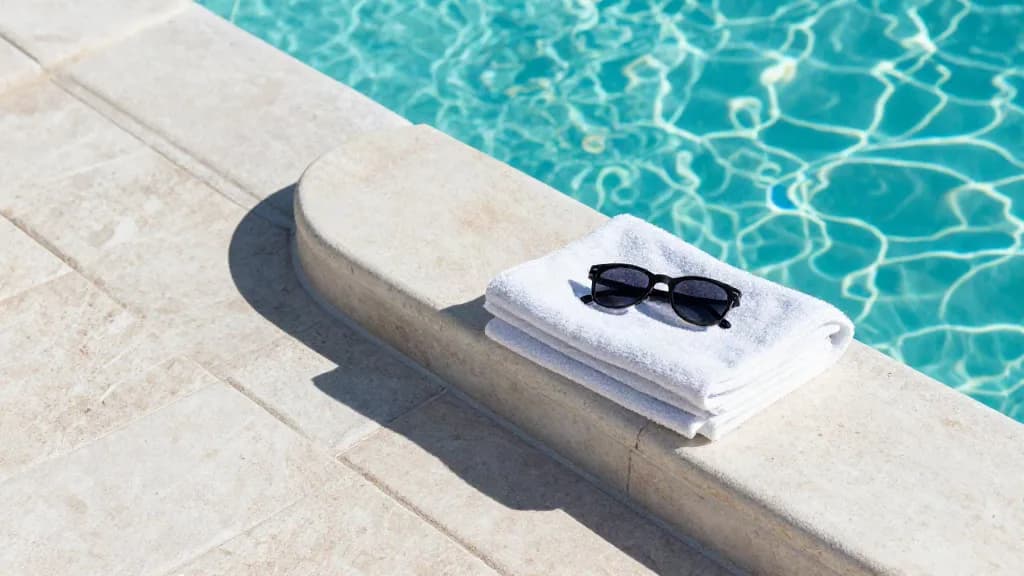 Light travertine pool deck and rounded coping with a towel and sunglasses by turquoise water